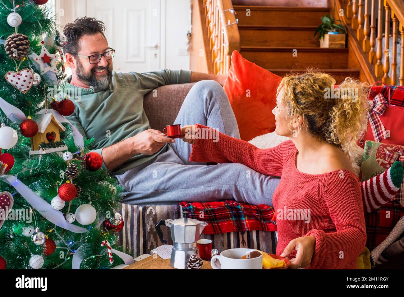 Un couple de jeunes adultes heureux à la maison appréciant le matin de noël en décembre prenant le café et le petit déjeuner ensemble. Homme sourire à une femme en vacances activité de loisirs à l'intérieur assis sur le canapé. Arbre de Noël Banque D'Images