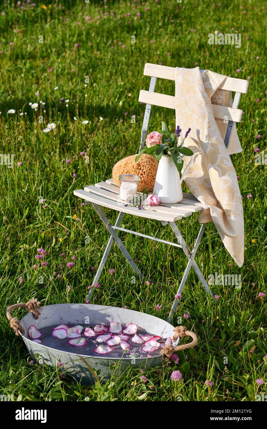 Marche pieds nus - des ustensiles tels que l'éponge naturelle, le sel, la lavande, les pétales pour le soin naturel des pieds sur une chaise de jardin blanche dans le vert, en face d'elle un bain de fleur dans un bain de zinc Banque D'Images