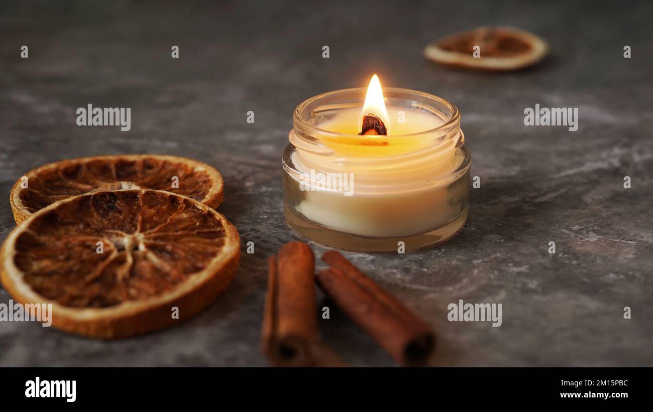 Bougie à la main brûlante avec mèche en bois dans un pot en verre blanc avec cannelle et oranges sèches à l'arrière-plan, décoration intérieure confortable d'hiver Banque D'Images