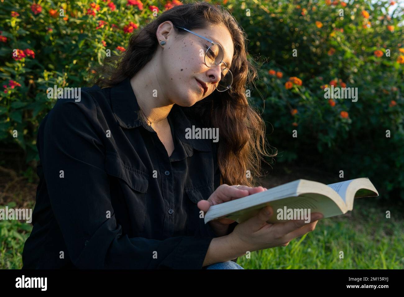 Femme pleine de style dans des vêtements décontractés et des lunettes de vue tout en lisant le livre dans le parc d'été le jour ensoleillé Banque D'Images