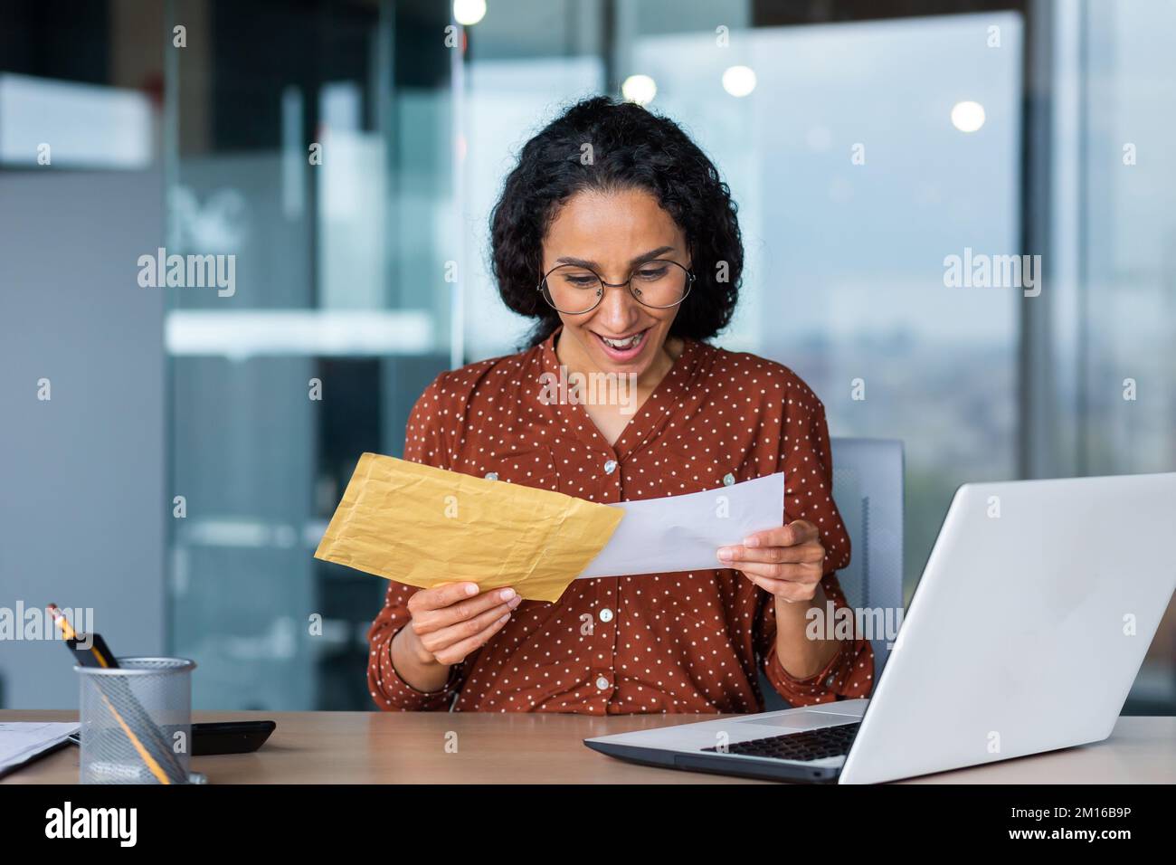 Une jeune femme latino-américaine heureuse a reçu une facture avec un salaire gagné, une récompense en espèces, de bonnes nouvelles. Il est assis au bureau, en tenant une enveloppe avec une lettre entre ses mains. Banque D'Images