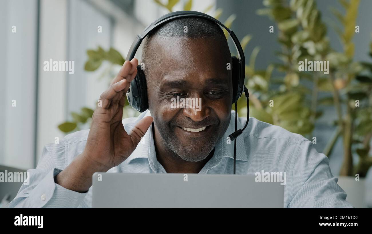 Un employé africain senior s'assoit au bureau, porte un casque avec microphone, se connecte à la vidéoconférence, permet de communiquer avec un ordinateur portable par webcam Banque D'Images