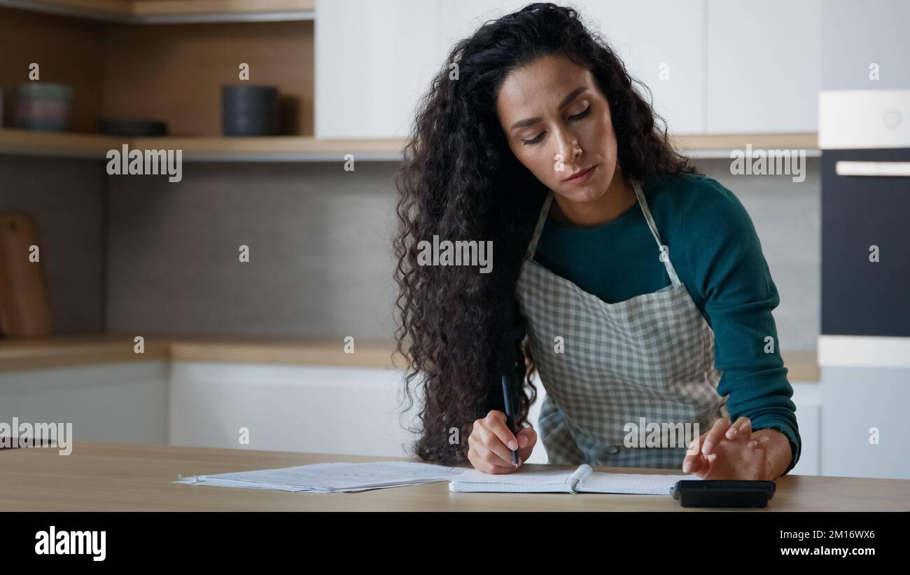 Concentrée femme hispanique femme jeune maman stand dans la cuisine moderne faire des notes à copybook écrire la recette d'épicerie liste calculer les services publics domestiques utilisation c Banque D'Images