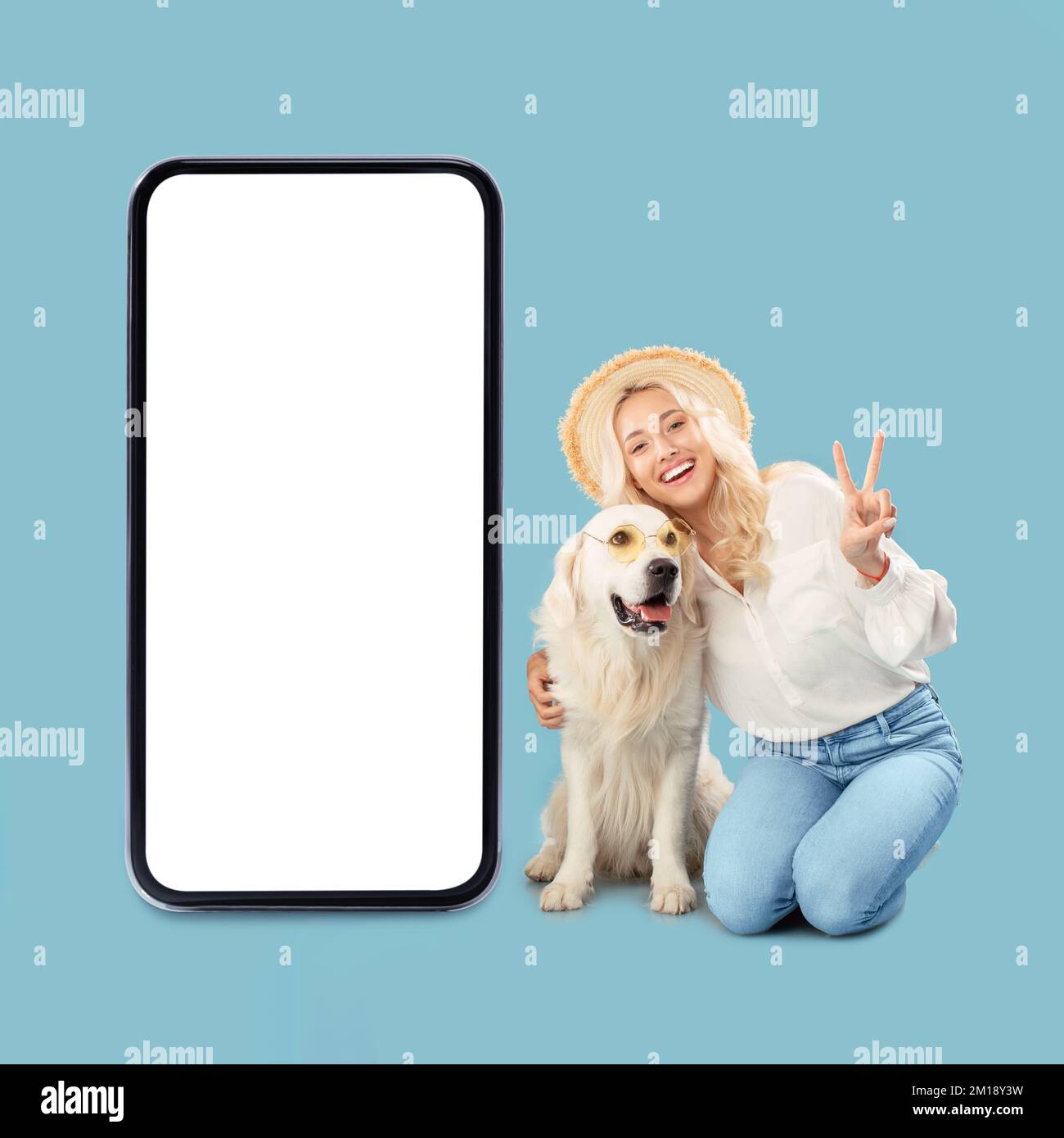 Une femme heureuse assise près de son chien du labrador et montrant un geste de paix, posant près d'un grand téléphone portable avec écran vide Banque D'Images