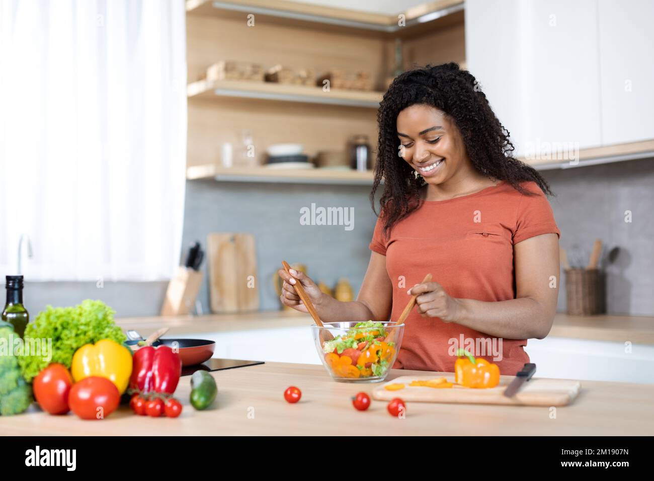 Mode de vie végétarien. Joyeuse jolie femme noire millénaire en t-shirt rouge faire de la salade, prépare le déjeuner pour la famille Banque D'Images