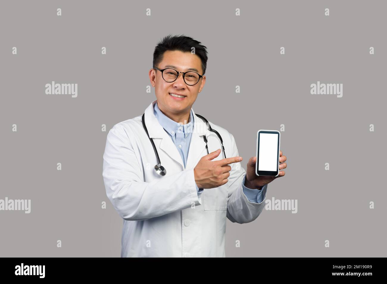 Souriant homme asiatique mûr en manteau blanc et lunettes pointe le doigt vers le smartphone avec écran vide Banque D'Images