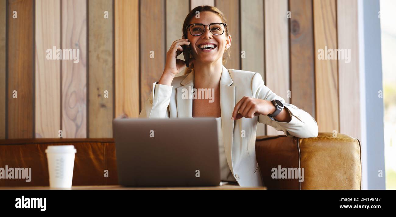 Une femme d'affaires assise dans un café, appelant au téléphone avec un sourire heureux sur son visage. Elle travaille à distance sur son ordinateur portable et son réseau wi Banque D'Images