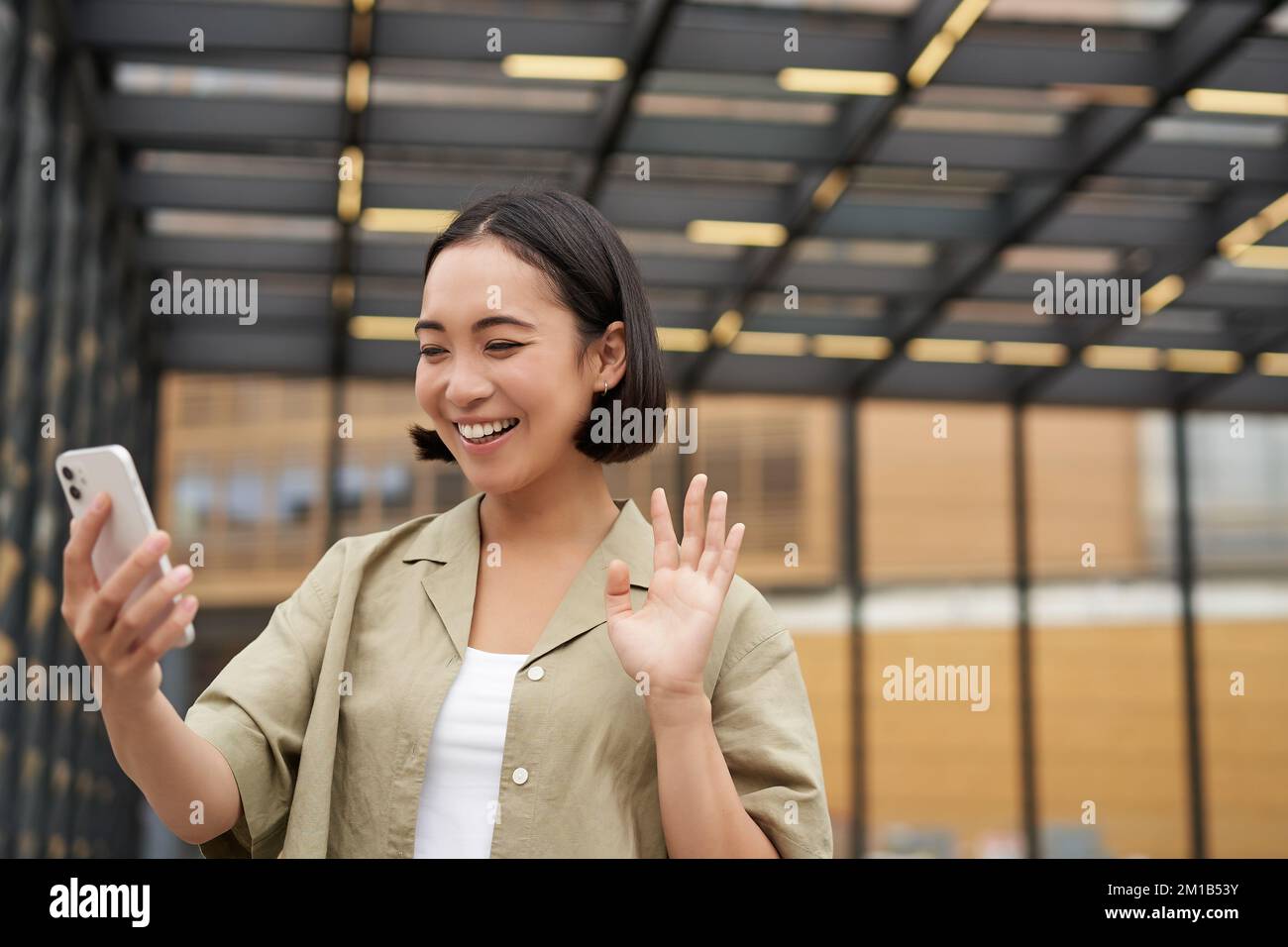 Une jeune fille asiatique se fait une joie de voir son appareil photo pour smartphone, dit bonjour à un ami lors d'un chat vidéo, appelant quelqu'un, debout dans la rue Banque D'Images