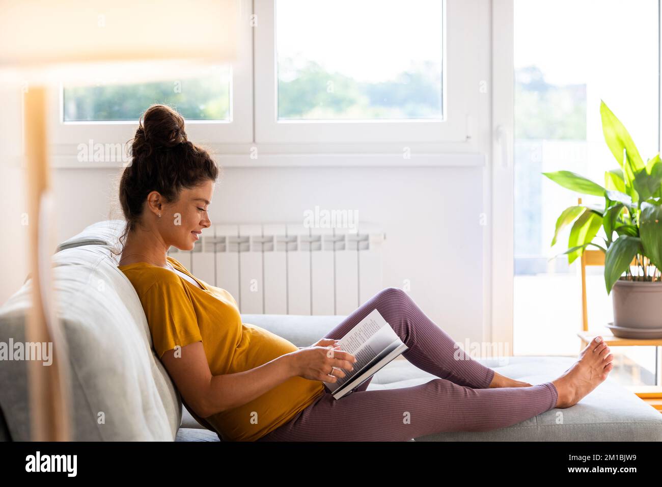 Jolie femme enceinte assise sur un canapé, repos et livre de lecture sur un canapé dans la salle de séjour Banque D'Images