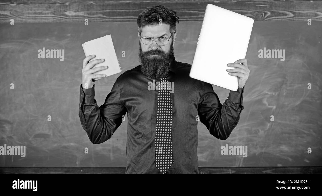 Numérique contre papier. Un hipster barbu pour enseignants tient un livre et un ordinateur portable. Que préférez-vous ? Enseignant choisissant l'approche moderne de l'enseignement. Choisissez droite Banque D'Images