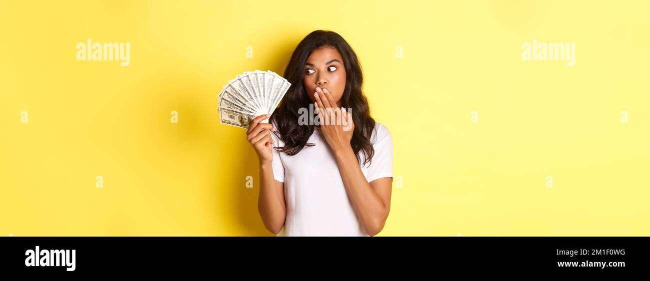 Image d'une fille afro-américaine heureuse et surprise, regardant une grosse somme d'argent avec un visage émerveillé, en regardant de l'argent liquide, debout sur jaune Banque D'Images