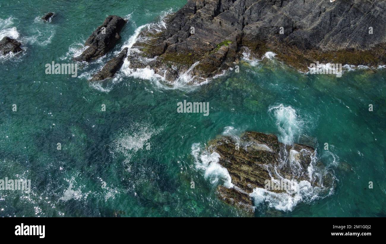 Rochers couverts de mousse noire au milieu des vagues turquoise de la mer celtique. Mousse de mer blanche sur les vagues. Eaux de l'océan Atlantique. Point de vue du drone. Banque D'Images