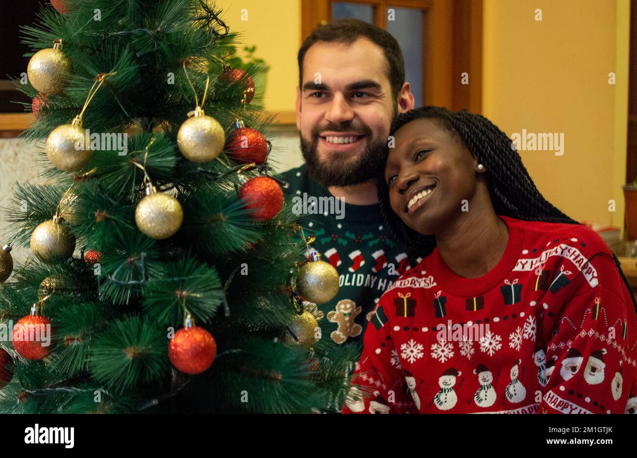 couple interracial appréciant noël embrassant devant un arbre décoré tout en portant des chandails de noël. Banque D'Images