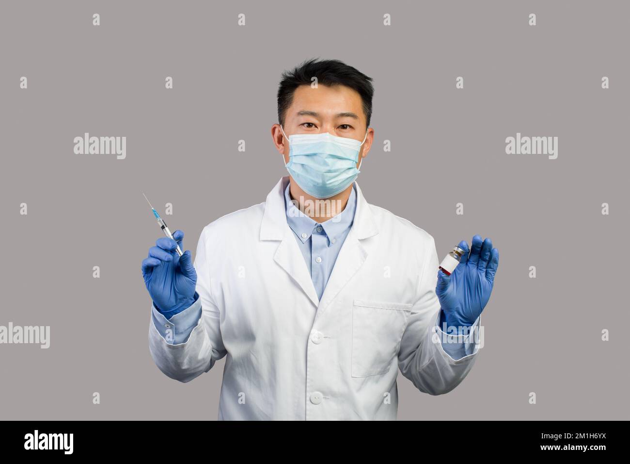 Homme chinois sérieux mature thérapeute en manteau blanc, masque de protection montre le vaccin et la seringue Banque D'Images