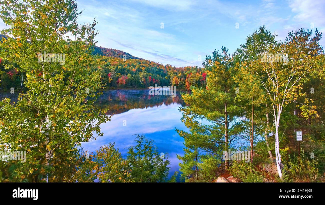 Vue à travers les arbres vue en hauteur sur le lac bleu du matin brumeux entouré par les montagnes d'automne Banque D'Images
