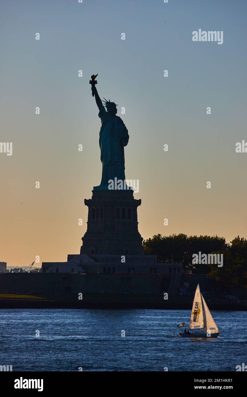 Silhouette de la Statue de la liberté avec un petit voilier qui passe à l'heure de la lumière et de l'or Banque D'Images