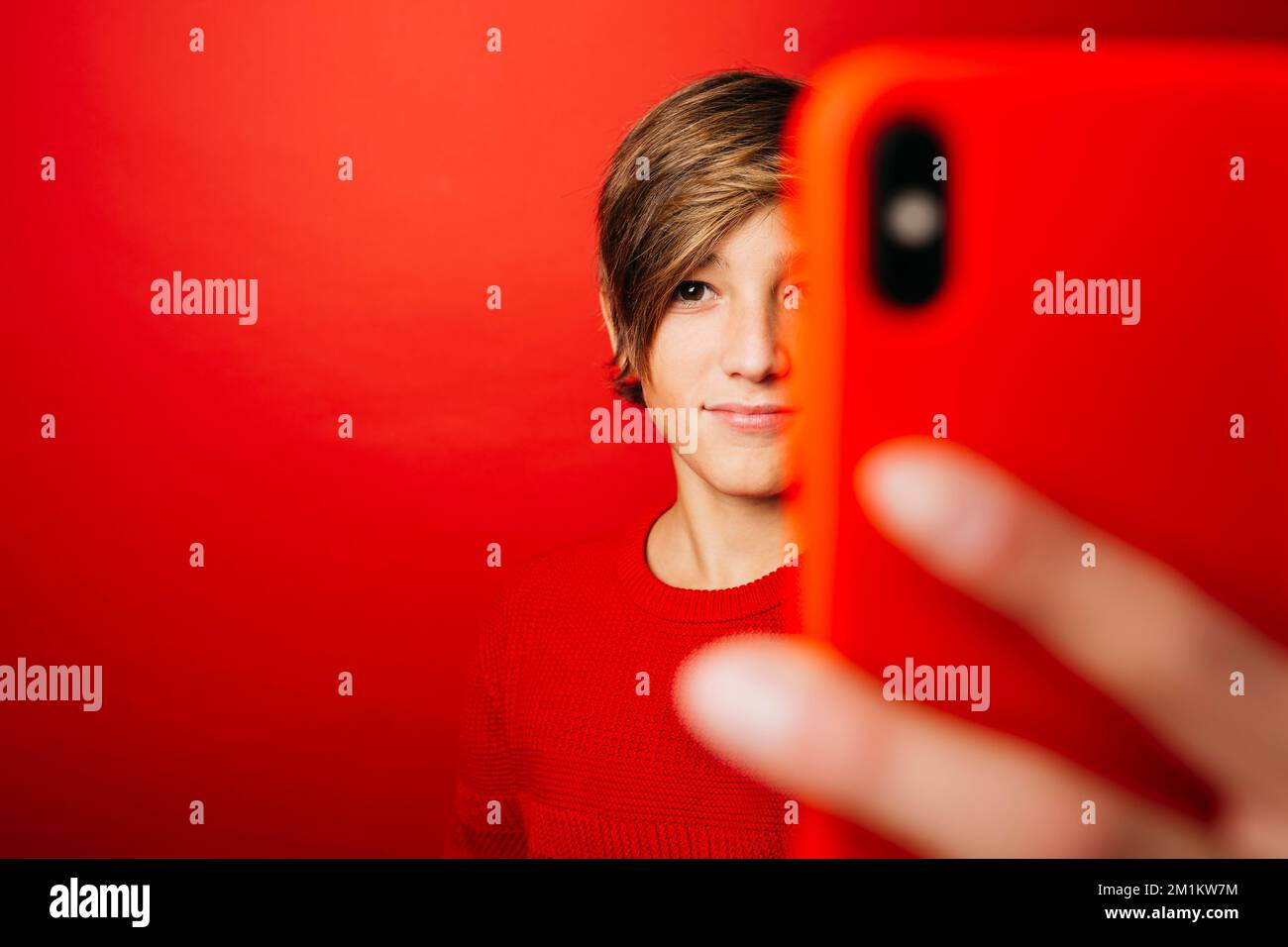 Pré-adolescent portant un chandail rouge sur fond rouge, prenant un selfie avec un smartphone rouge Banque D'Images