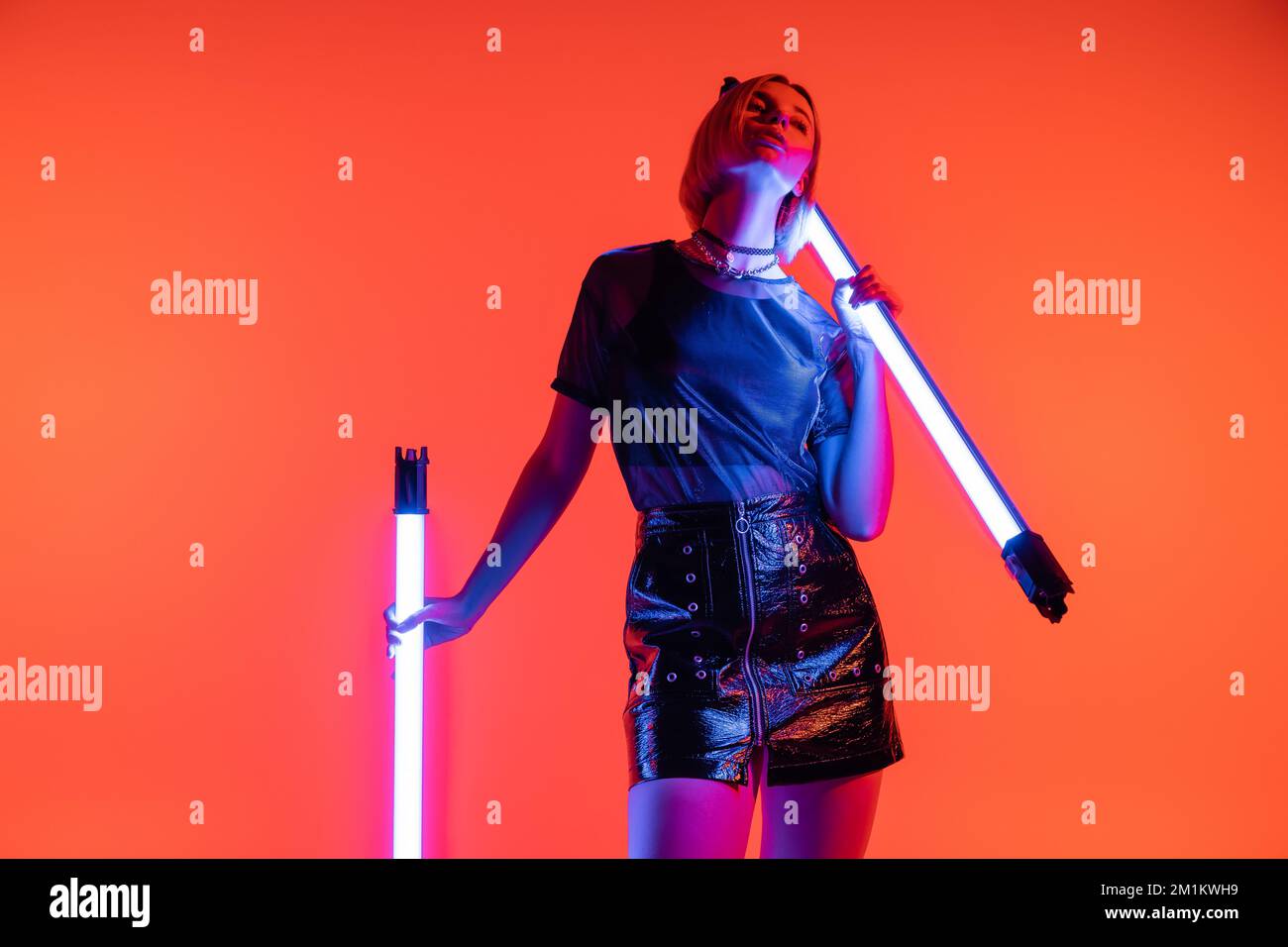 jeune femme en t-shirt à la mode et mini jupe en cuir posant avec des lampes au néon sur fond rouge corail, image de stock Banque D'Images