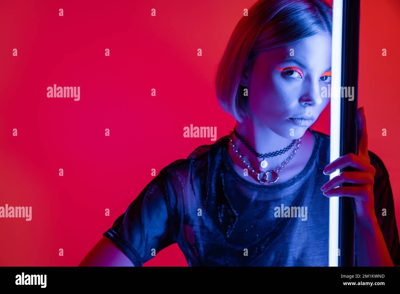 femme élégante en maquillage lumineux et colliers regardant l'appareil photo dans la lumière bleue de la lampe néon sur fond rouge carmin, image de stock Banque D'Images