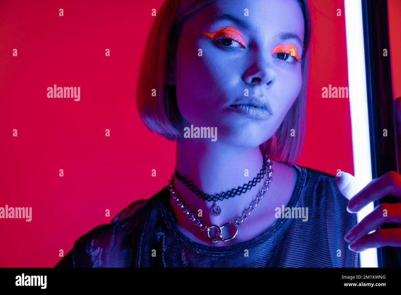 femme tendance dans le maquillage lumineux et colliers regardant la caméra dans la lumière bleue de la lampe néon sur fond rouge carmin, image de stock Banque D'Images