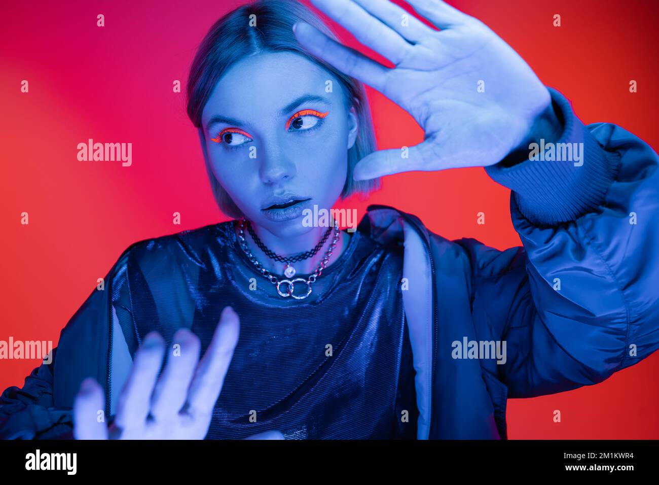 jeune femme en maquillage de néon regardant loin en posant avec des mains tendues sur fond rouge de corail, image de stock Banque D'Images