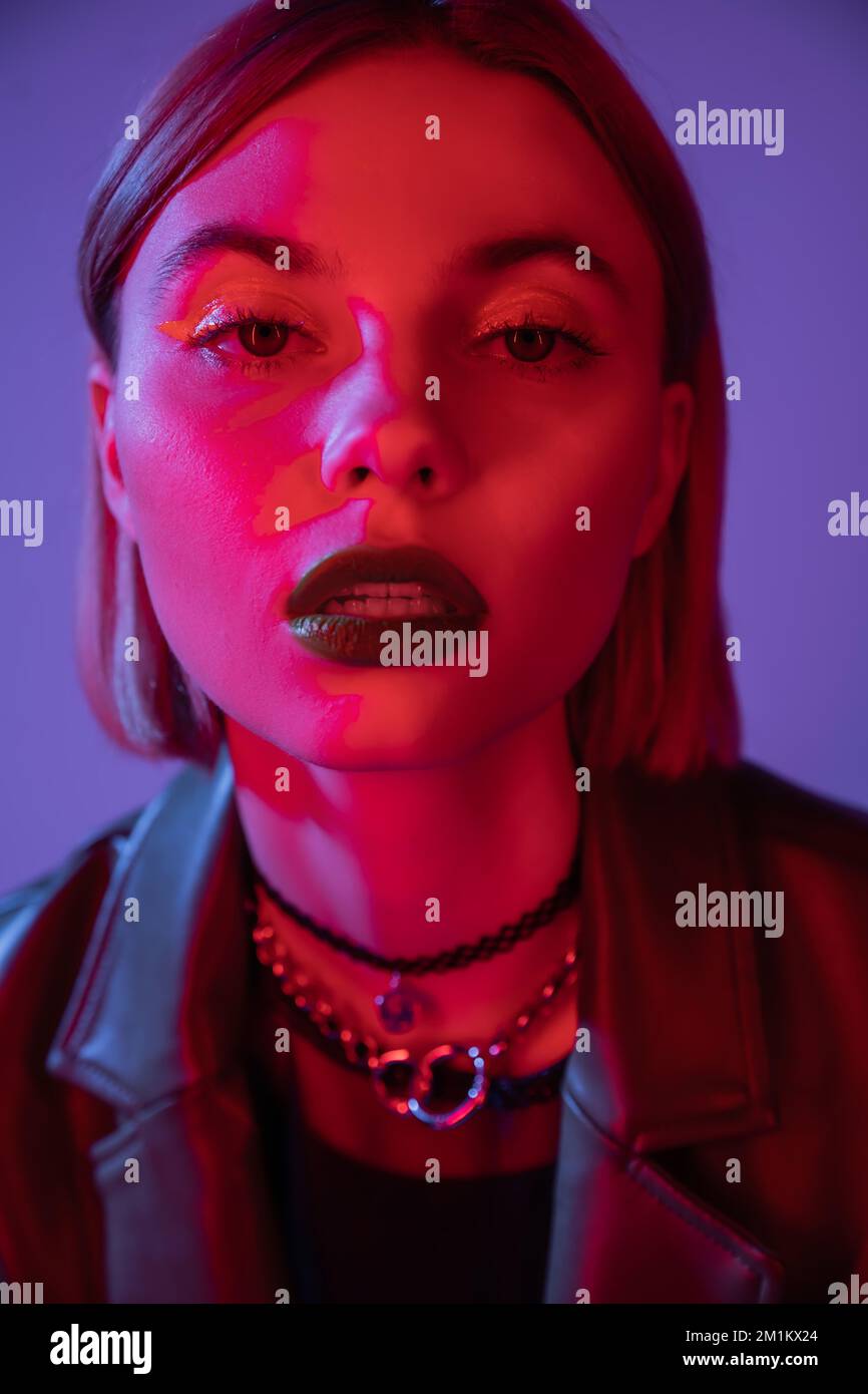 portrait d'une femme en maquillage brillant regardant l'appareil photo dans la lumière rouge isolée sur violet, image de stock Banque D'Images