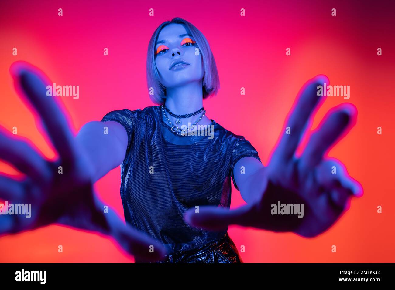vue à angle bas de la femme avec maquillage lumineux et mains étirées dans la lumière bleue néon sur fond corail et rose, image de stock Banque D'Images
