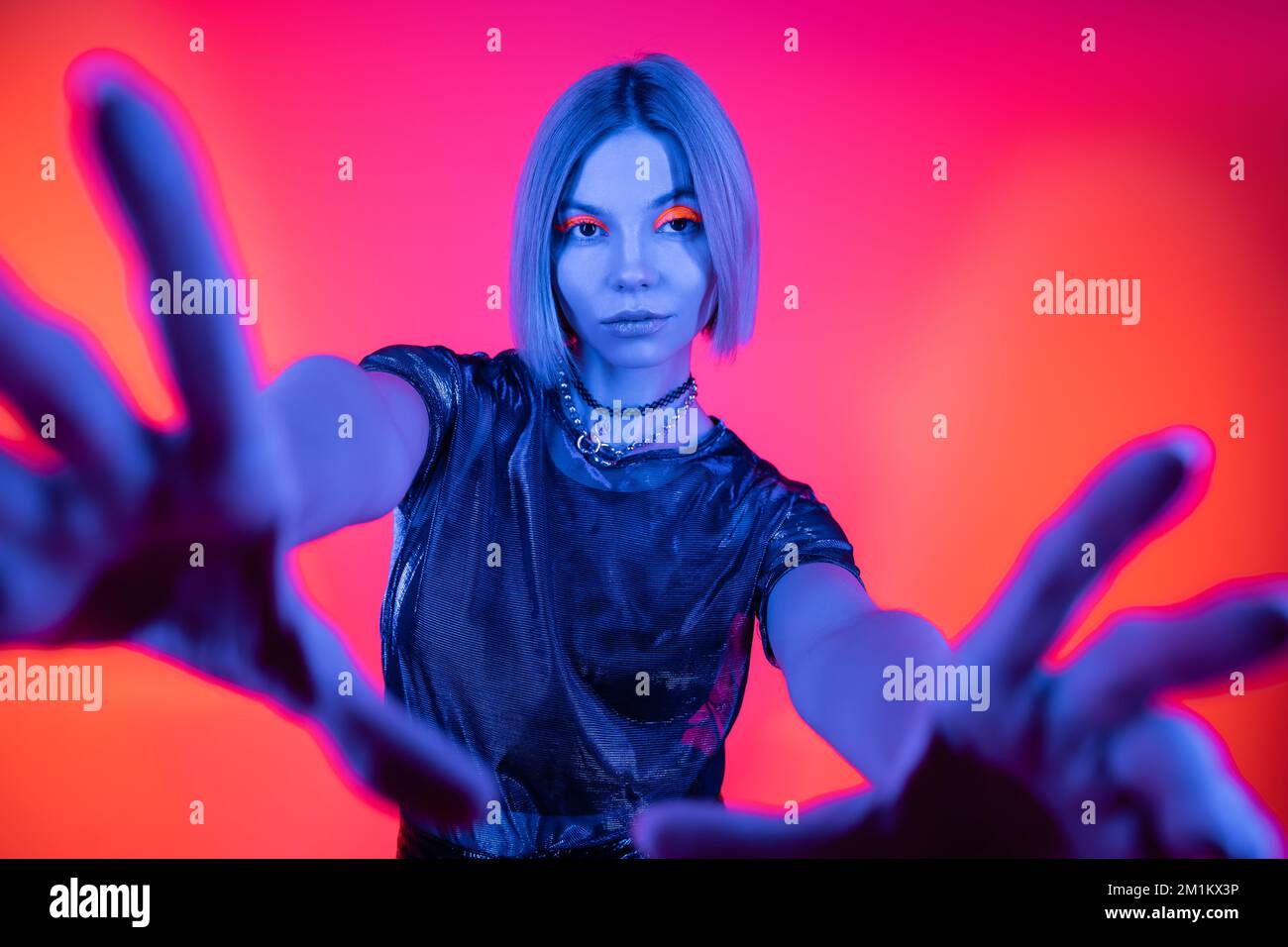 femme élégante avec des mains étirées regardant la caméra dans la lumière bleue néon sur fond corail et rose, image de stock Banque D'Images