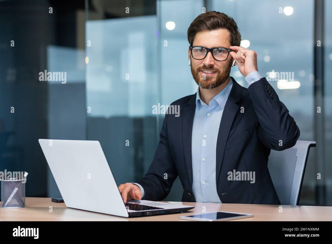 Portrait d'un jeune homme d'affaires prospère, avocat, défenseur juridique. Regarde en toute confiance l'appareil photo, tient des lunettes, rit, s'assoit au bureau à la table, travaille à l'ordinateur portable. Banque D'Images