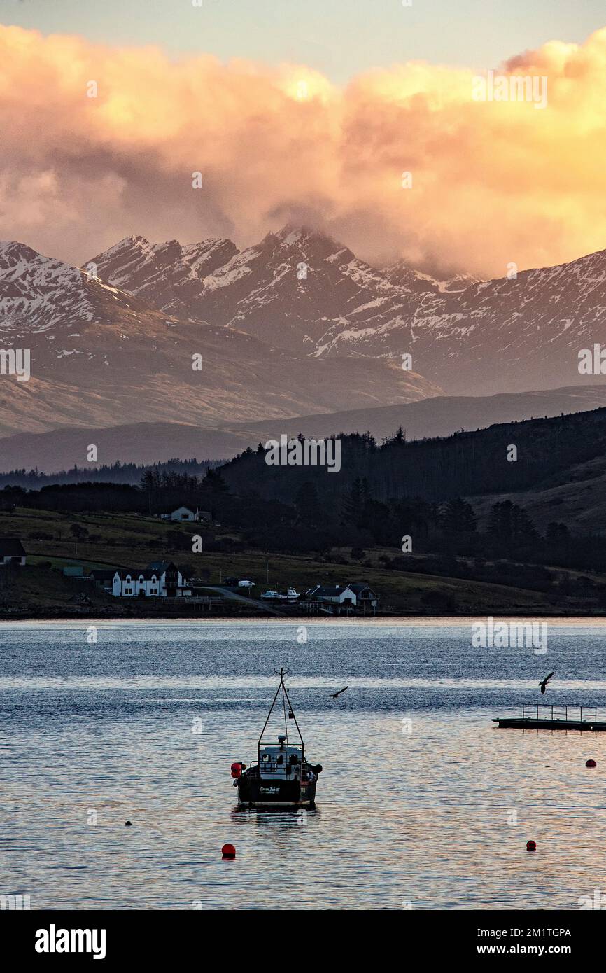 Vue sur le port de Portree vers Camustianavaig avec un petit bateau de pêche en premier plan, des maisons périphériques et la lumière du soleil orange sur les nuages A. Banque D'Images