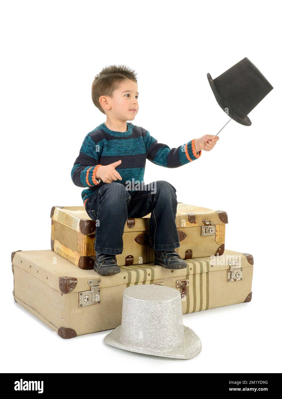 Garçon isolé en Jean et pull-over rayé assis sur quelques vieilles valises joue avec une baguette et un chapeau de dessus. Banque D'Images