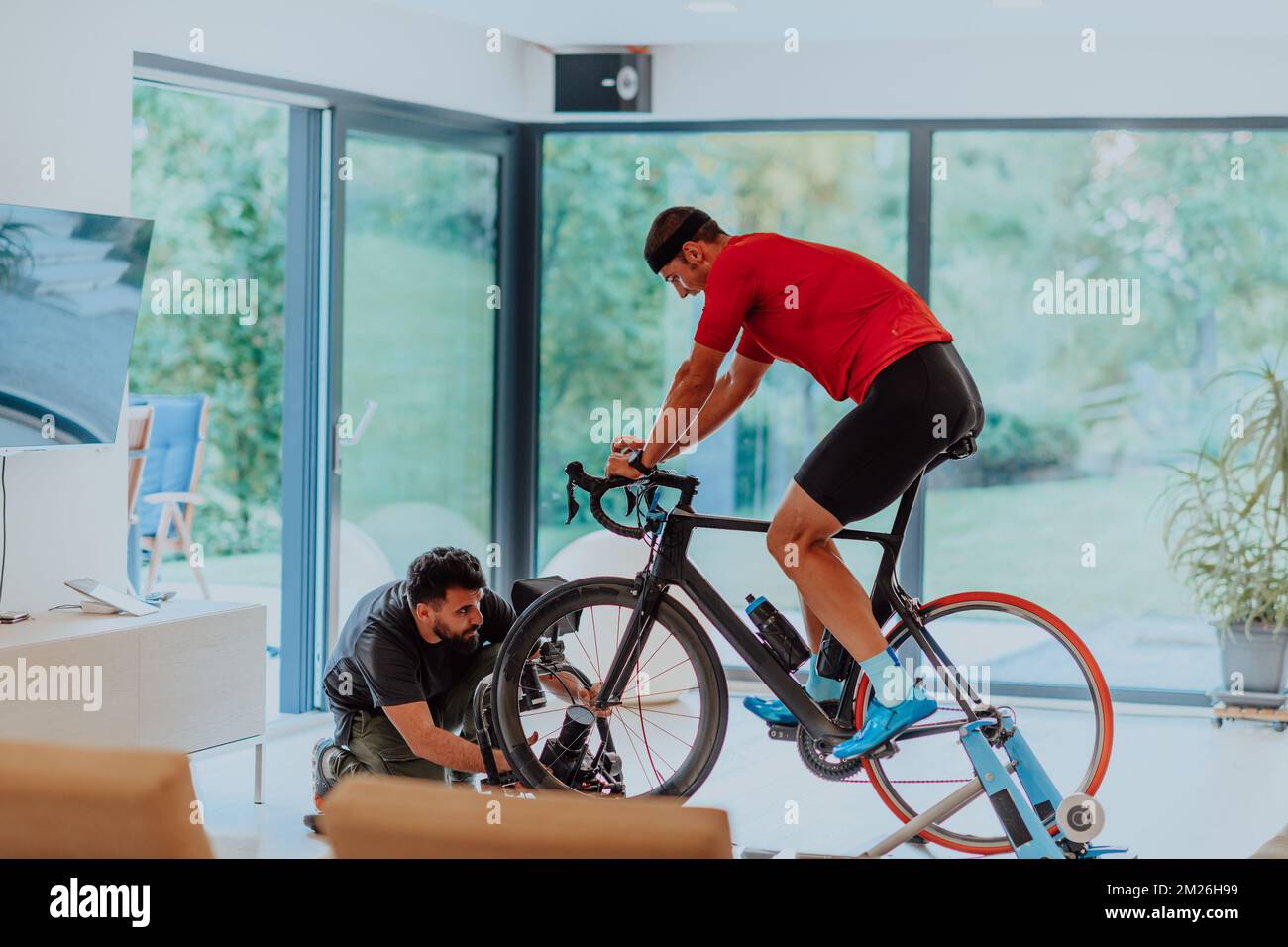 Un caméraman filmant un athlète à vélo de triathlon sur une machine de simulation dans un salon moderne. Formation en cas de pandémie. Banque D'Images