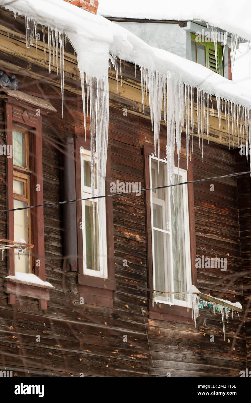 Plusieurs glaçons sont suspendus au bord du toit, en hiver ou au printemps. Mur en bois d'une ancienne maison en bois avec fenêtres. De grandes cascades de glaçons dans des rangées lisses et magnifiques. Jour d'hiver nuageux, lumière douce. Banque D'Images