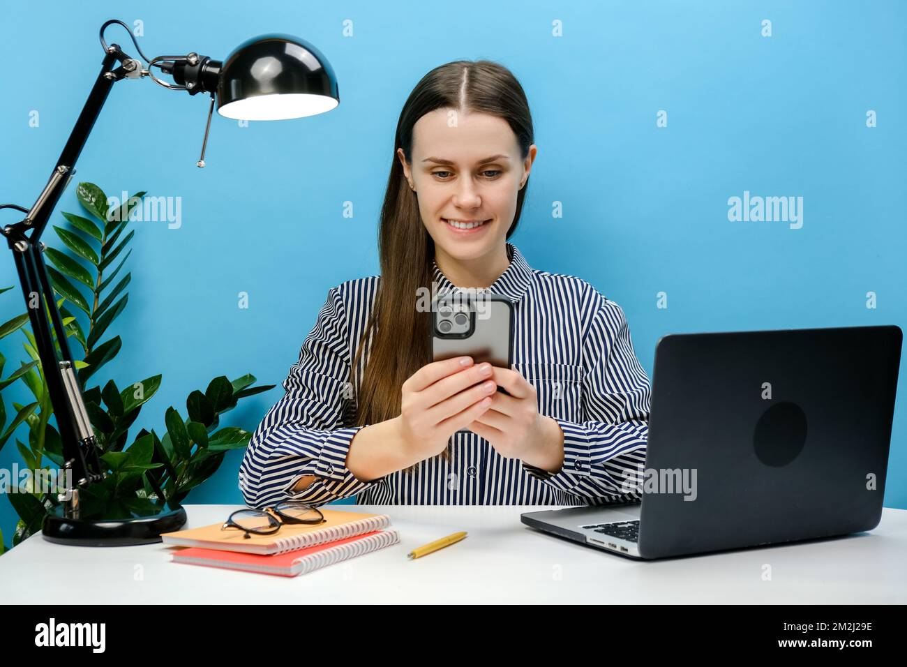 Souriant belle employée réussie jeune femme s'assoir au bureau blanc avec pc portable tenant mobile téléphone mobile surfer sur Internet, p Banque D'Images