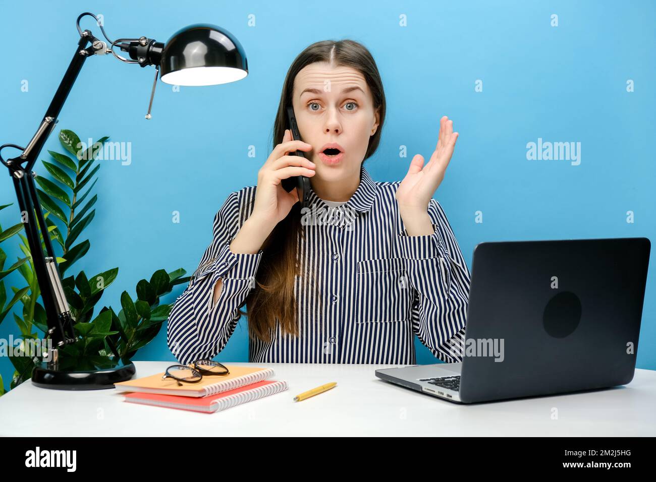 Portrait d'un employé confus choqué femme d'affaires assis au bureau blanc avec un ordinateur portable parlant sur le téléphone mobile se répandre les mains, posant iso Banque D'Images