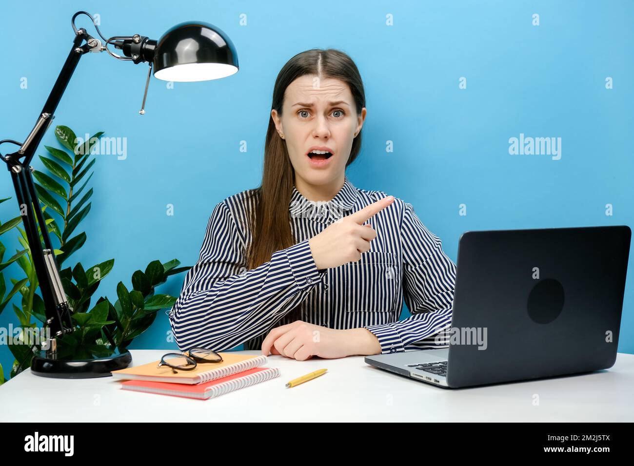 Une jeune femme d'affaires en colère en chemise s'assoit au bureau avec un pc portable pointant l'index de côté, isolé sur un mur bleu. Le bus de la réussite Banque D'Images