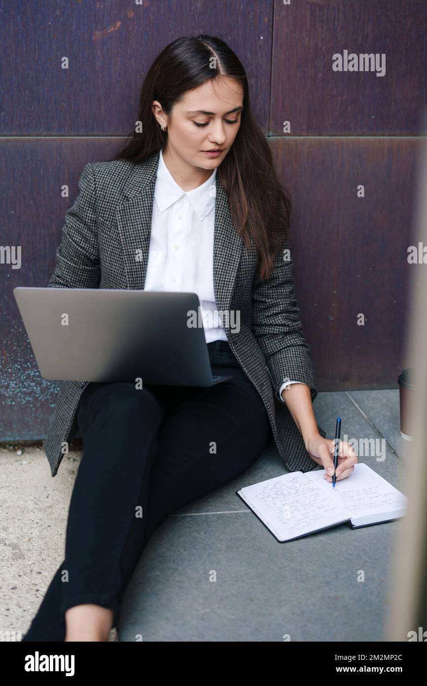 Belle femme caucasienne utilisant un ordinateur portable tout en écrivant dans un ordinateur portable, assis à l'extérieur sur le sol. Communication avec les professionnels. Communication moderne Banque D'Images