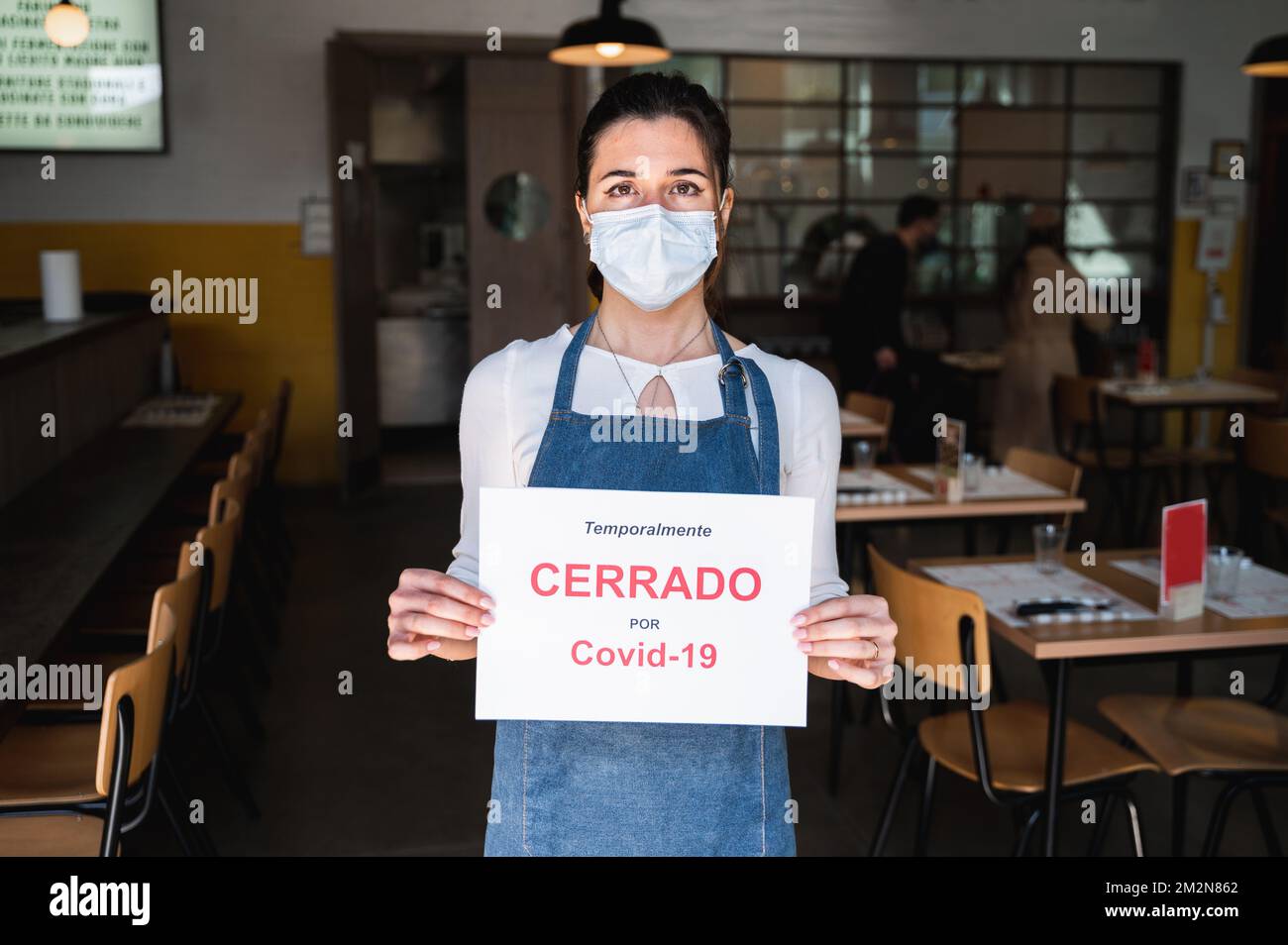 Femme propriétaire d'affaires avec masque facial et tablier tenant une pancarte avec l'inscription fermée en raison de covid en espagnol. Banque D'Images