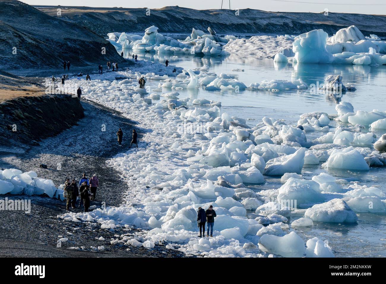 On voit des gens marcher près des zones de glace sur la rive du lac Jökulsárlón. Jökulsárlón est un lac situé dans le sud de l'Islande, avec une superficie de 20 km2 et une profondeur de plus de 200 mètres. Jusqu'à il y a moins de 100 ans, le glacier de Breiðamerkurjökull (qui fait partie du glacier Vatnajökull) s'étendait même au-delà du périphérique. En raison de la hausse des températures, le glacier a reculé, créant cette impressionnante lagune glaciaire. Banque D'Images