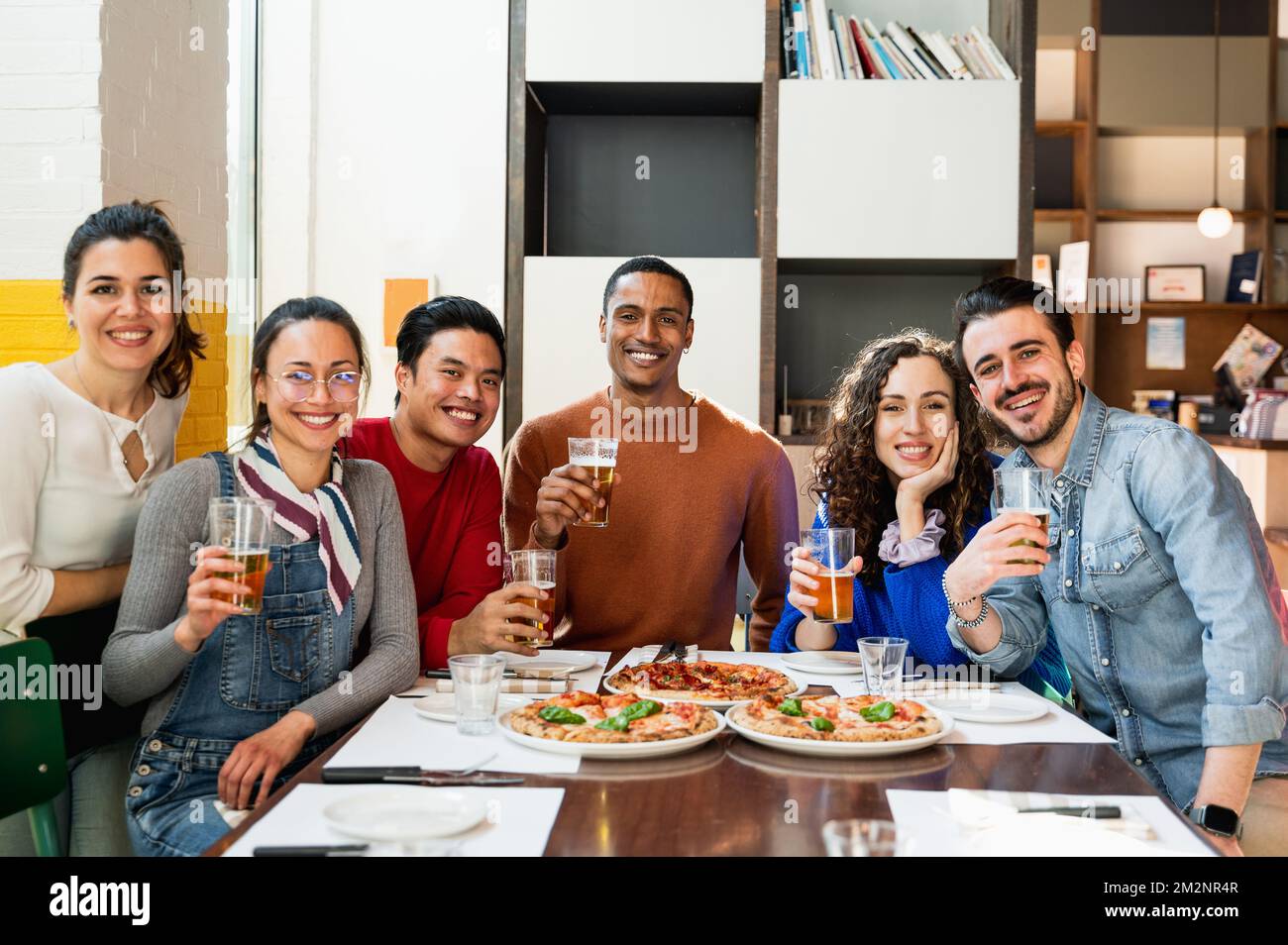 Portrait d'un groupe multiethnique de jeunes amis assis à table, mangeant de la pizza et buvant de la bière, posant à la caméra avec des expressions souriantes. Banque D'Images
