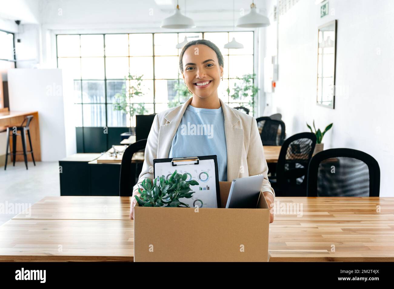 Joyeuse latino ou femme d'affaires brésilienne, dans des vêtements élégants, debout dans son nouveau bureau de travail, tenant une boîte avec des articles de travail dans ses mains, a obtenu un nouveau travail, regardant la caméra, souriant heureux Banque D'Images