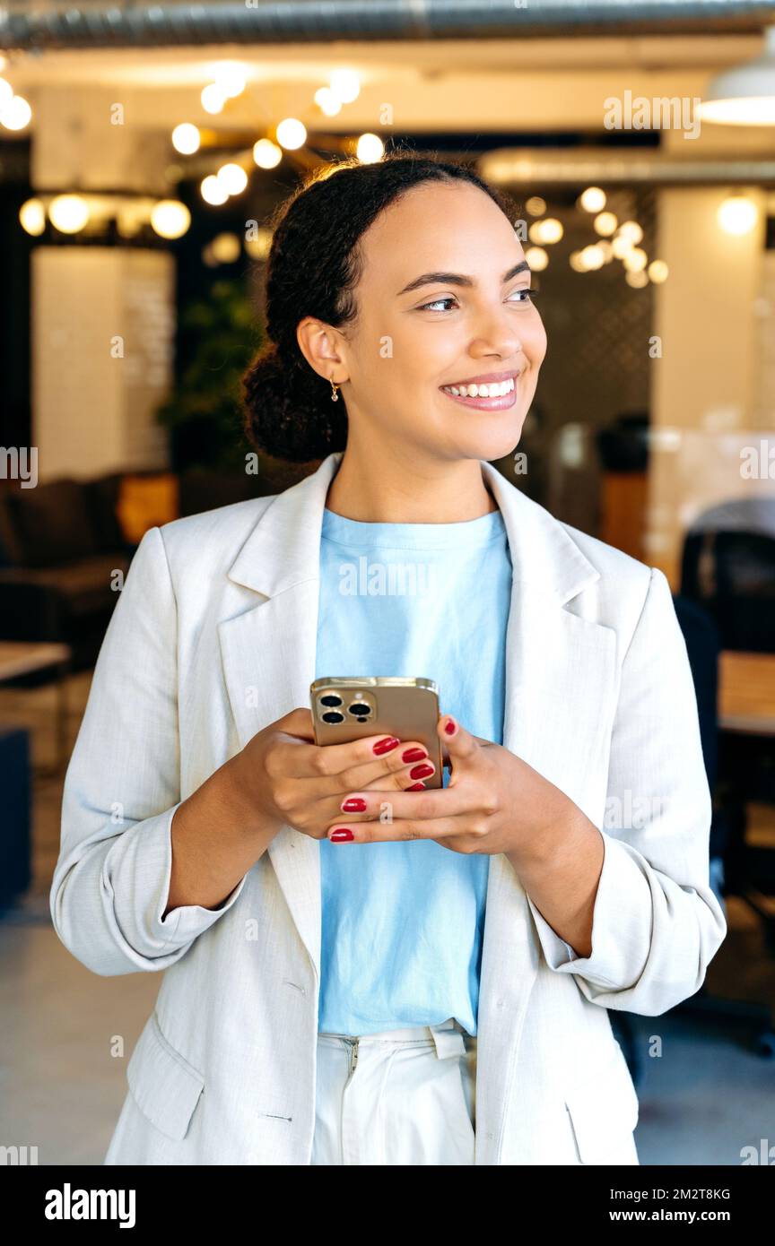 Messagerie en ligne avec un collègue, un client, un ami. Positive mixte race femme, directeur d'entreprise, stand dans le bureau moderne, utilise son smartphone, messagerie dans les médias sociaux, répond email, taper du texte, sourires Banque D'Images