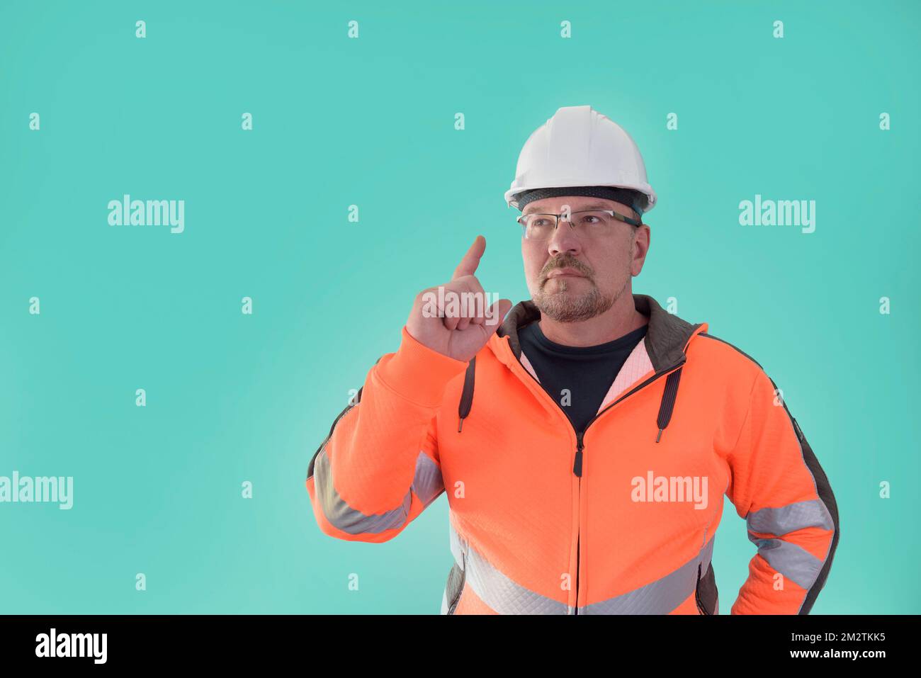 Un homme de construction, pensant, doigt vers le haut, dans des vêtements de travail et un casque de construction, sur un fond vert clair. Banque D'Images