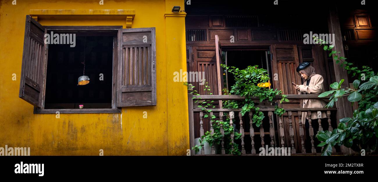 Une femme se tient sur un balcon à côté d'une fenêtre ouverte avec une bougie et une lampe suspendue dans un bâtiment jaune à Hoi an. Banque D'Images