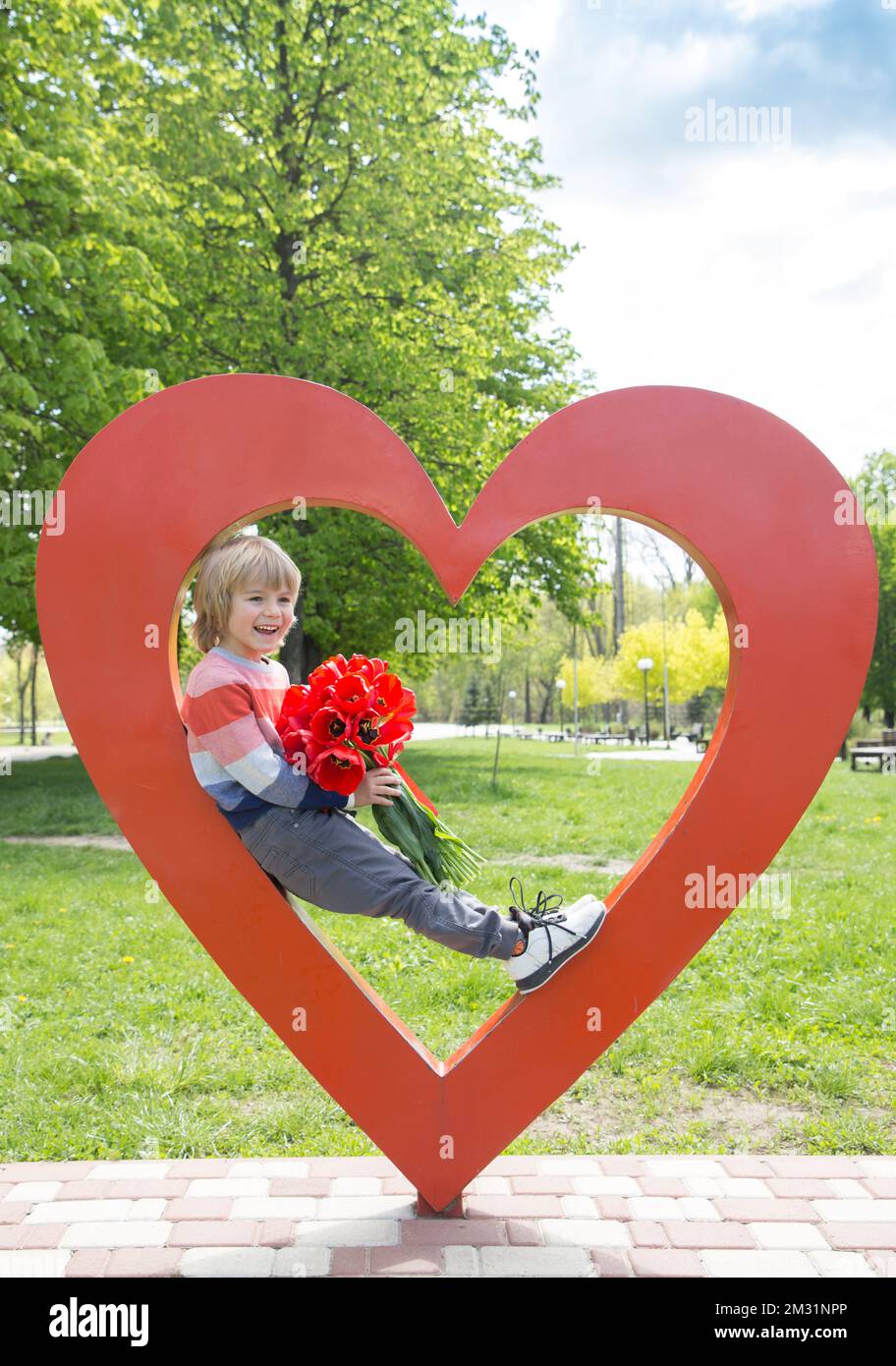 un garçon joyeux et émotionnel de 5-6 ans, avec un grand bouquet de tulipes rouges, se trouve à l'intérieur d'un grand coeur rouge. Fête des mères, fête de la Saint-Valentin. Cadeau de fleur avec l Banque D'Images