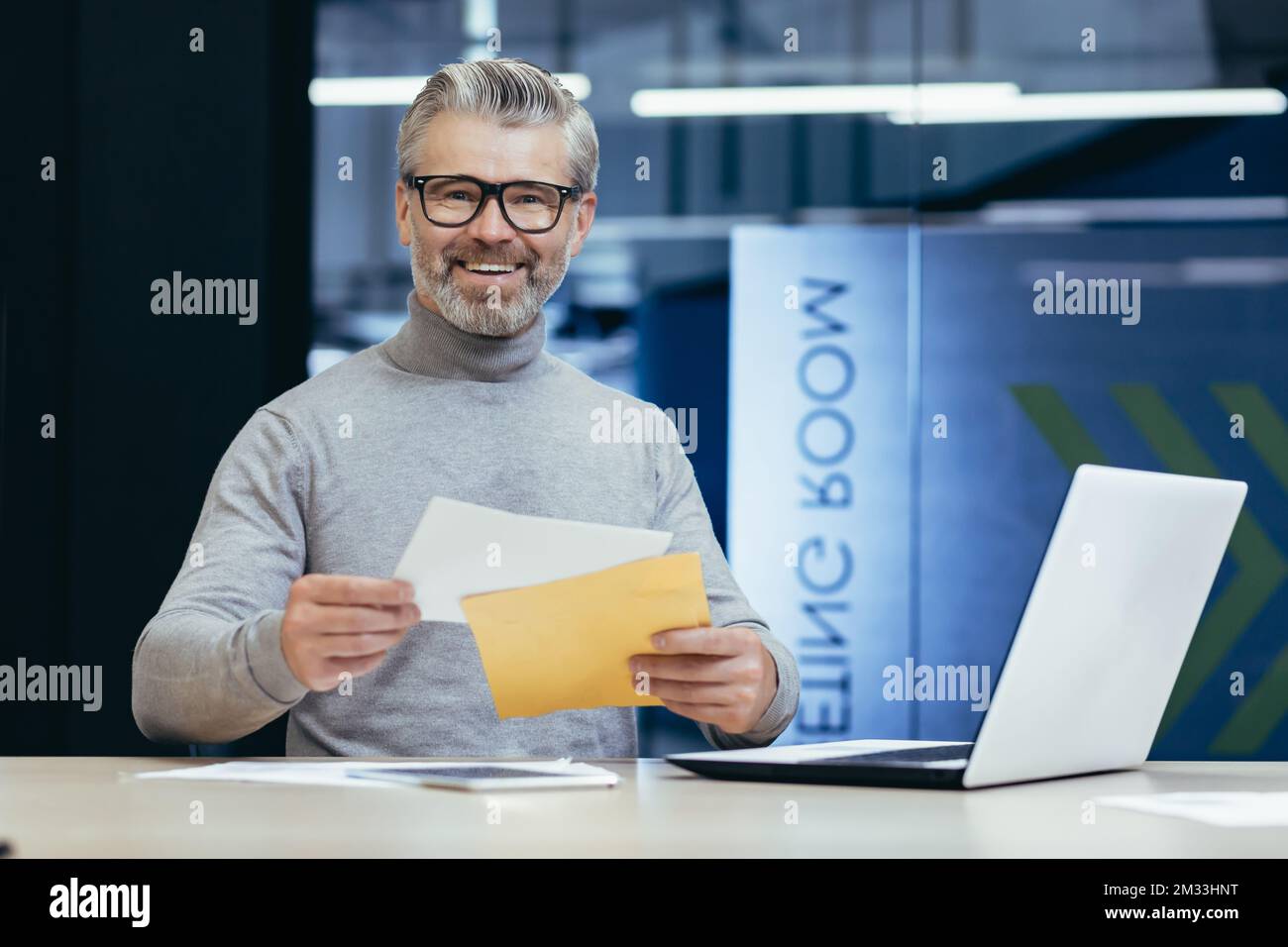 Heureux homme senior assis dans le bureau à la table et tenant une enveloppe dans sa poche. J'ai reçu de bonnes nouvelles, un prêt. J'ai fait un contrat. Il regarde la caméra, sourit. Banque D'Images