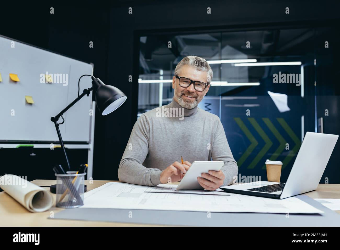 Homme aux cheveux gris, architecte, concepteur, ingénieur. Assis au bureau avec un ordinateur portable et de la paperasse. Il tient un ordinateur portable avec un crayon dans les mains. Il regarde la caméra et sourit. Banque D'Images