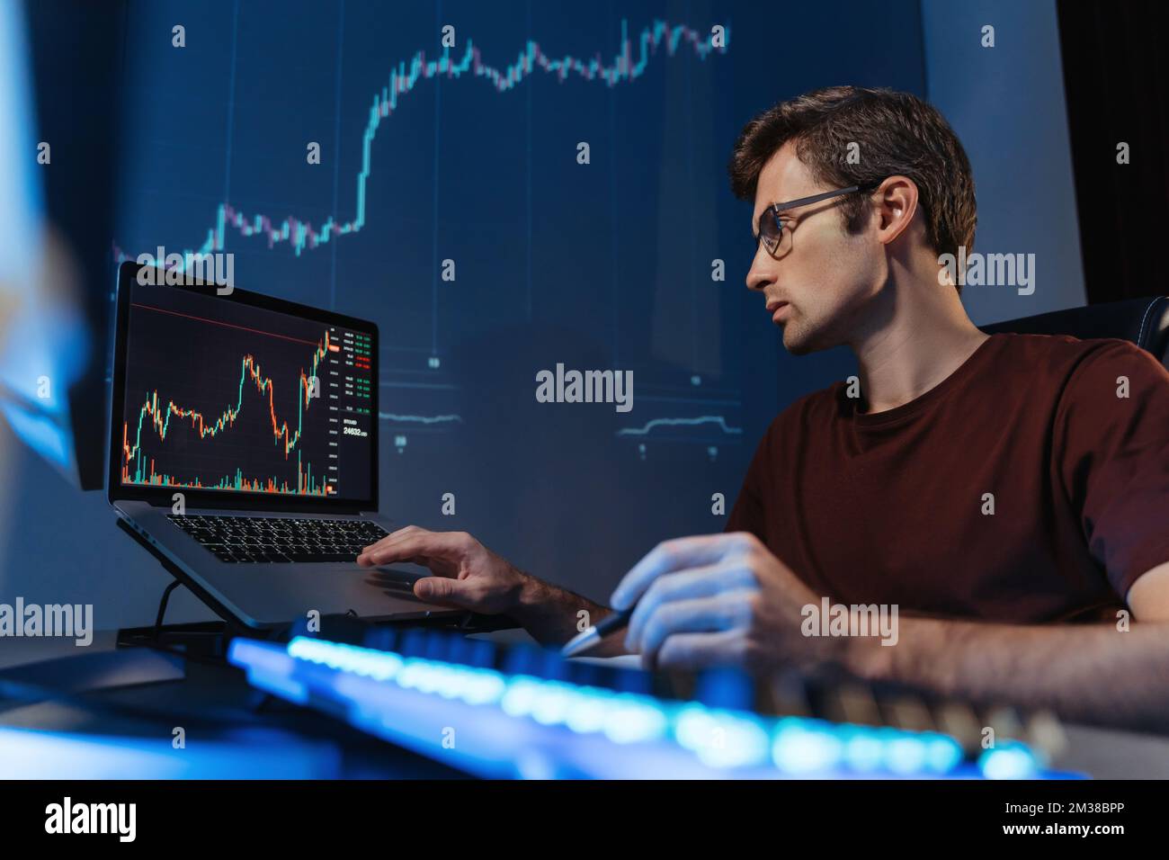 Expert en crypto financière des lunettes travaillant tard dans la nuit en ligne sur ordinateur portable au bureau à domicile, analysant le diagramme de chandelier regardant l'écran du moniteur, sur le mur numérique avec le diagramme Banque D'Images