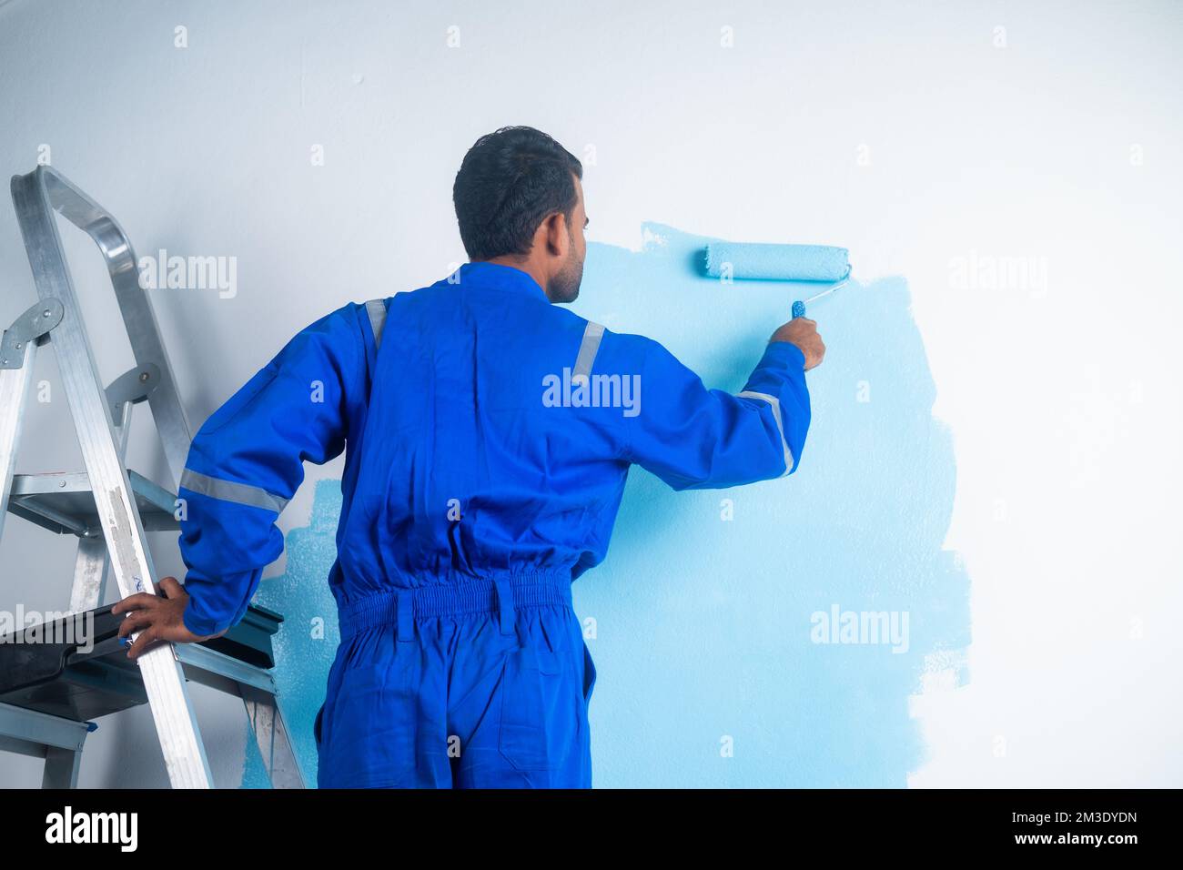 Vue arrière de peintre faisant des travaux de peinture sur le mur avec le rouleau en se tenant sur l'échelle - concept de - le travail dur, le travail de col bleu et Banque D'Images
