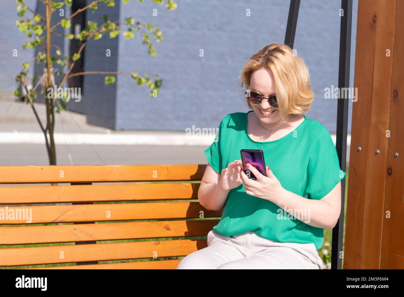 Portrait d'une fille étudiante souriante dans des lunettes de soleil,  tenant un smartphone dans ses mains, assis sur un banc dans un parc de  ville.Une fille étudiant remotel, en utilisant Photo Stock -